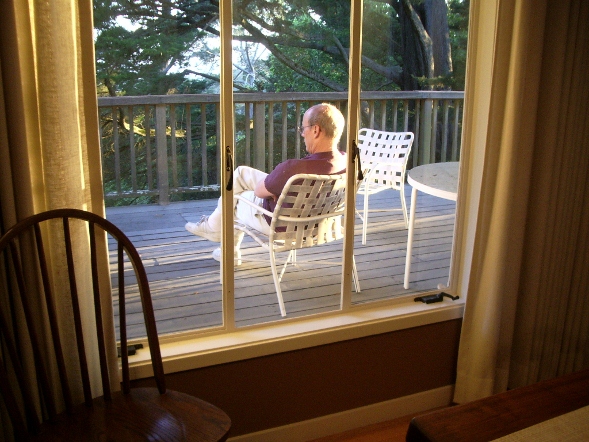 Jon Newhall sitting on our deck quietly, seen through a window, with trees. Photo by Barbara Falconer Newhall