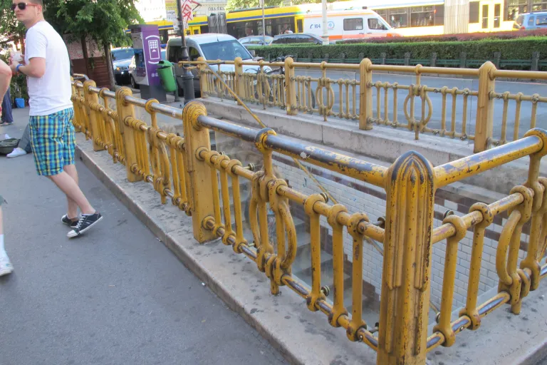 A battered balustrade and subway tiles on a sidewalk entrance to a Budapest subway. Yellow paint is peeling away. Photo by Barbara Newhall